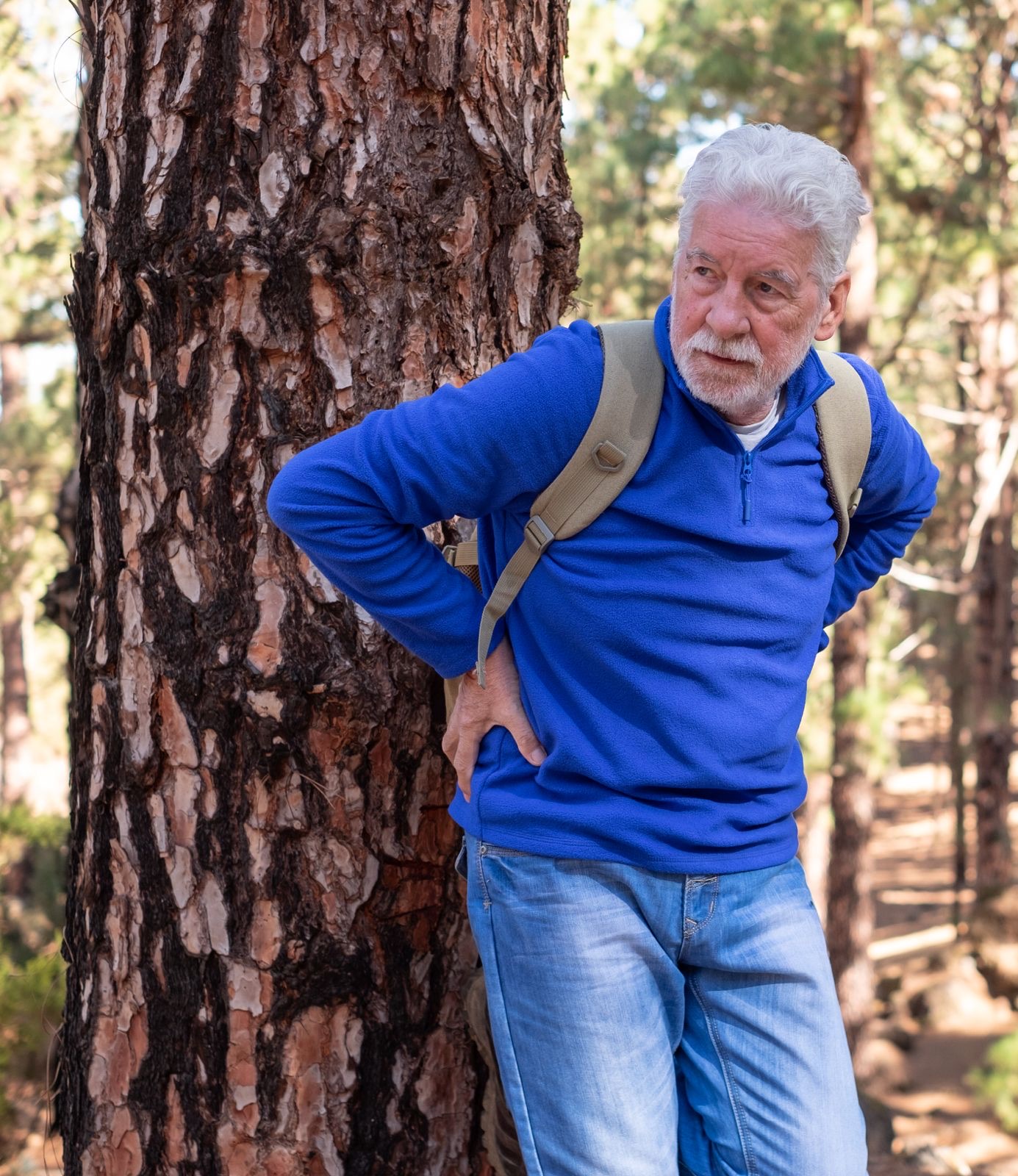 A man on a forest trail, stopped with a hand on his lower back